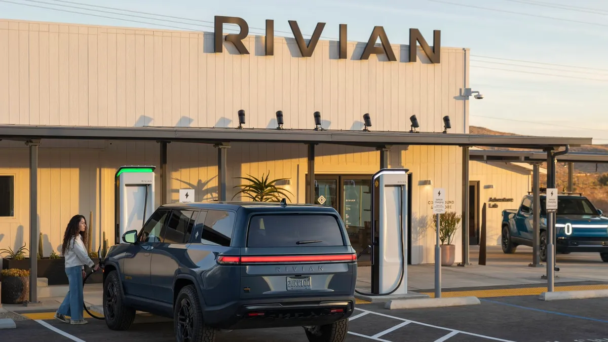 A person charges a blue Rivian R1S SUV outdoors with the sun shining at the company's Joshua Tree Charging Outpost in California