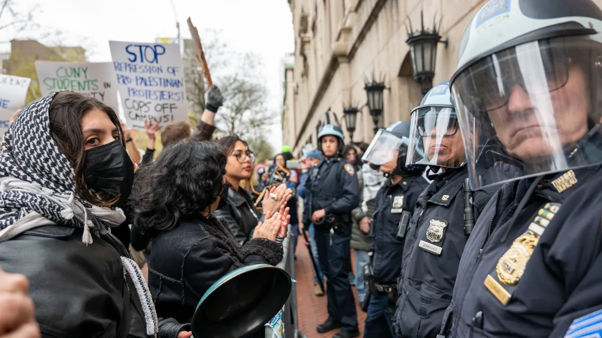 A line of young protestors face a line of police officers in riot gear.