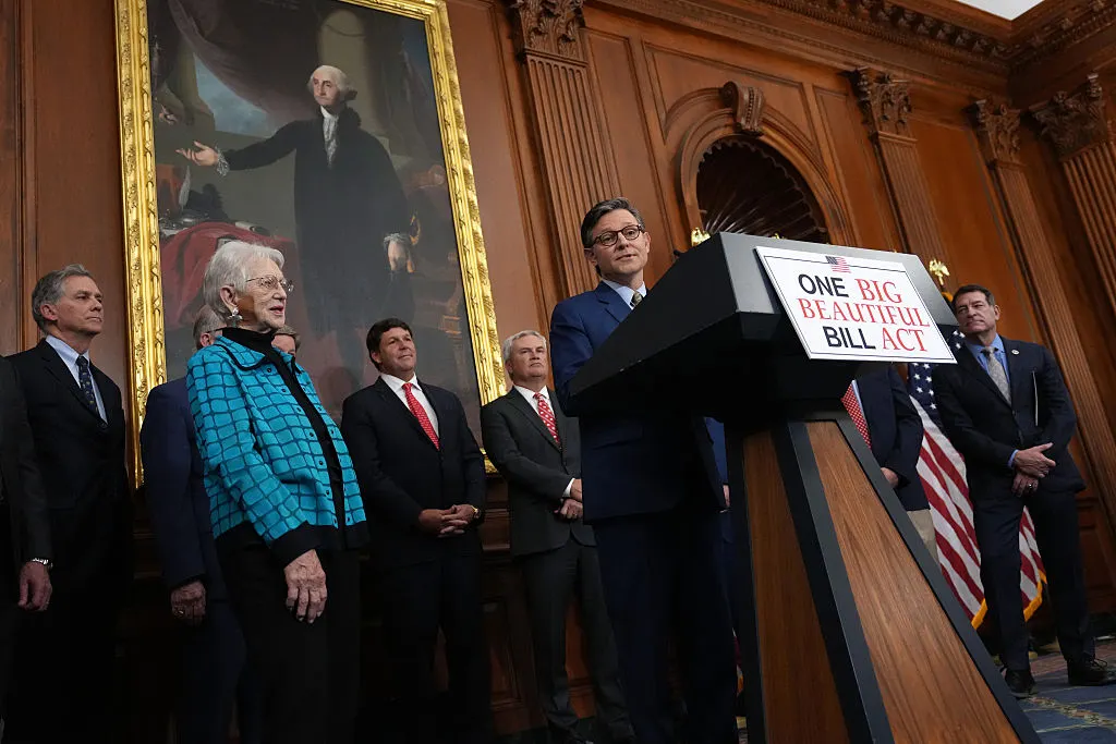 One person is standing behind a podium with a sign that reads "One Big Beautiful Bill Act." A handful of other people are standing around and behind the person and they are in a room with wood paneling.