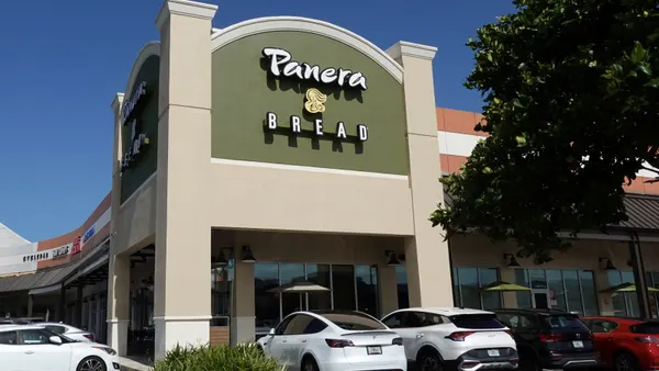 A photogrpah of a beige and green corner building wih several cars parked in front. The facade has a white sign that says Panera Bread.