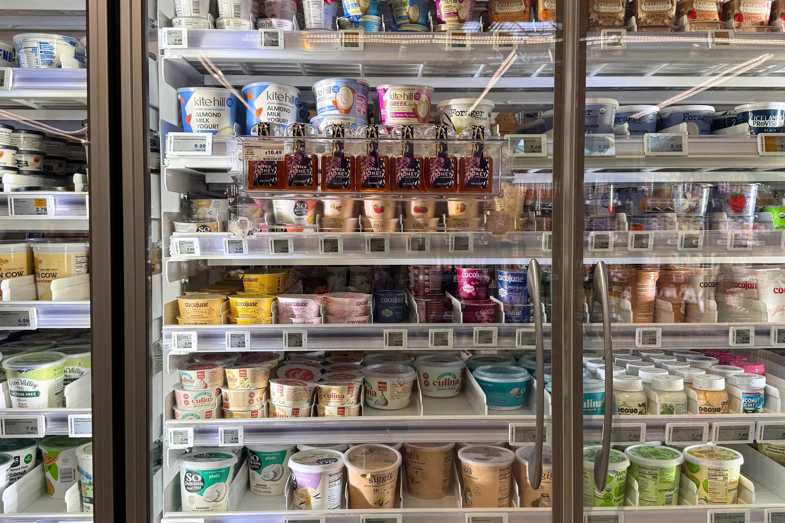 Glass-door grocery cooler stocked with assorted yogurt and dairy-free alternatives in neatly arranged rows, with visible price tags and brands like Kite Hill, Forager, Siggi’s, and Cocojune.