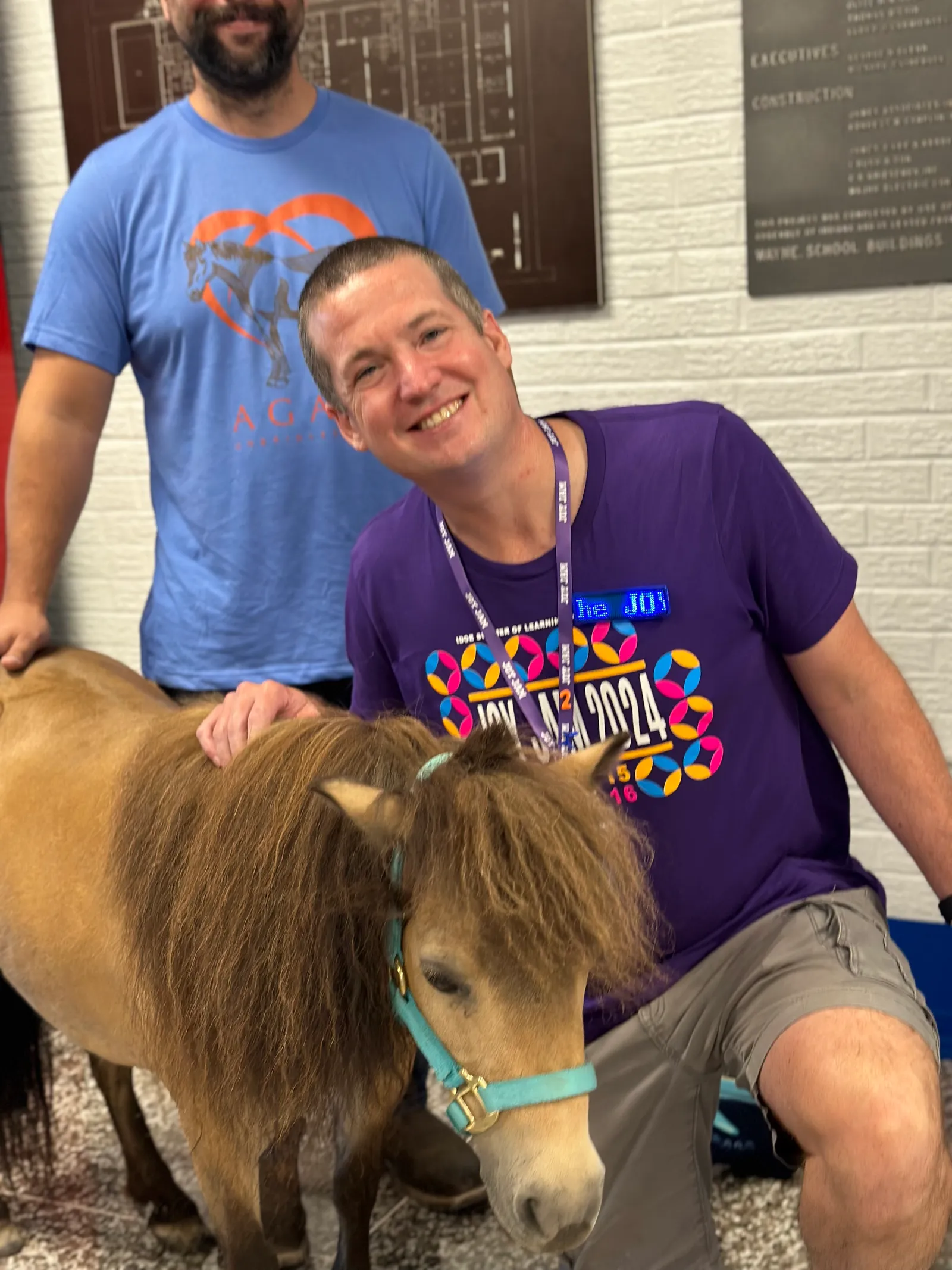 A man school superintendent kneels to pose with a mini horse at a school district celebration.