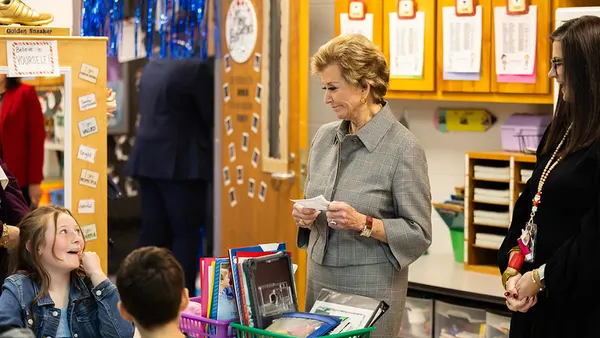 Two adults are standing in a classroom. A student is seated at a desk looking up at one adult.