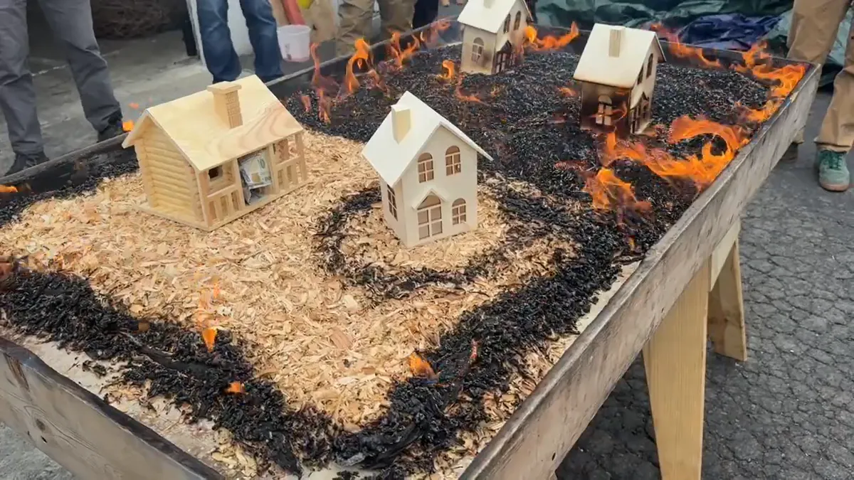 A tabletop with a small wooden house with a $100 bill inside, surrouned by woodchips on fire and a group of people looking on.