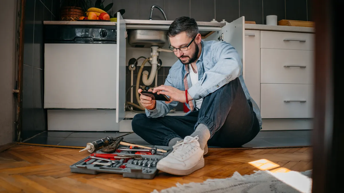 A man looks at his phone while repairing a sink.