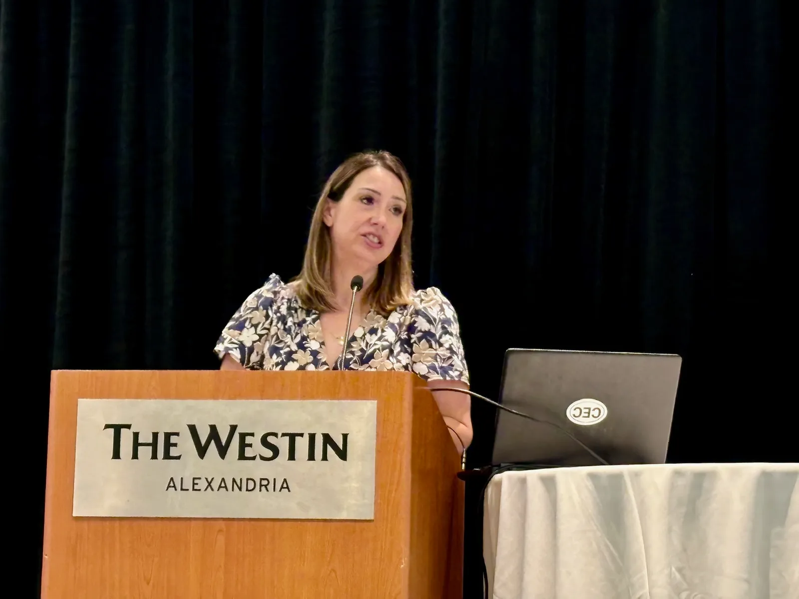 A person stands at a wooden podium with a microphone that has a sign reading &quot;The Westin Alexandria.&quot; To the person&#x27;s left is a table with a white tablecloth. on top of the table is a laptop.