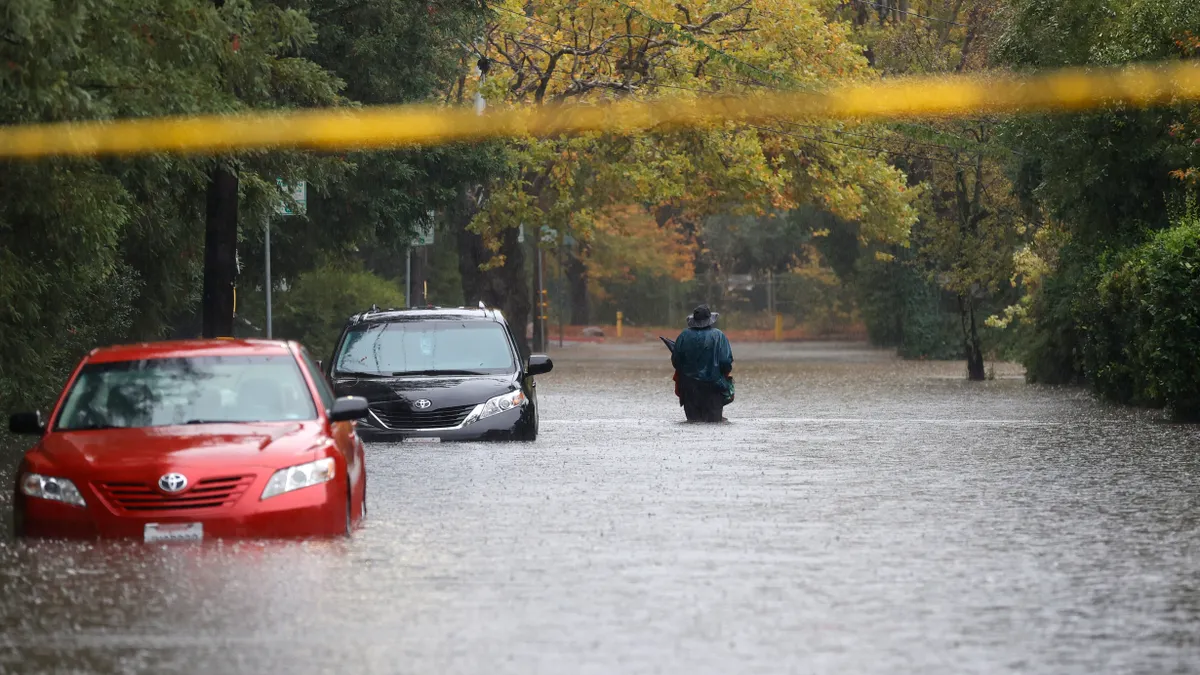 A red car and black car are half-submerged on a flooded road bordered by trees as a person in rainwear walks with his back to the camera.