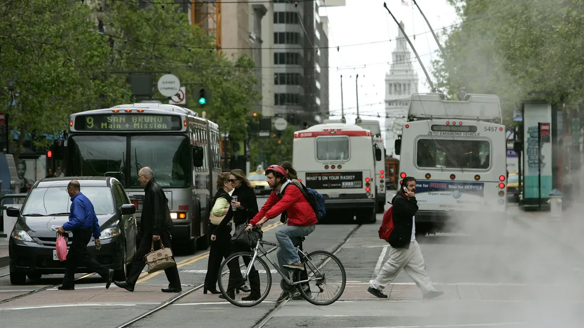 A bicyclist rides along Market Street near pedestrians crossing the street in San Francisco, California. Three buses and a car are visible.