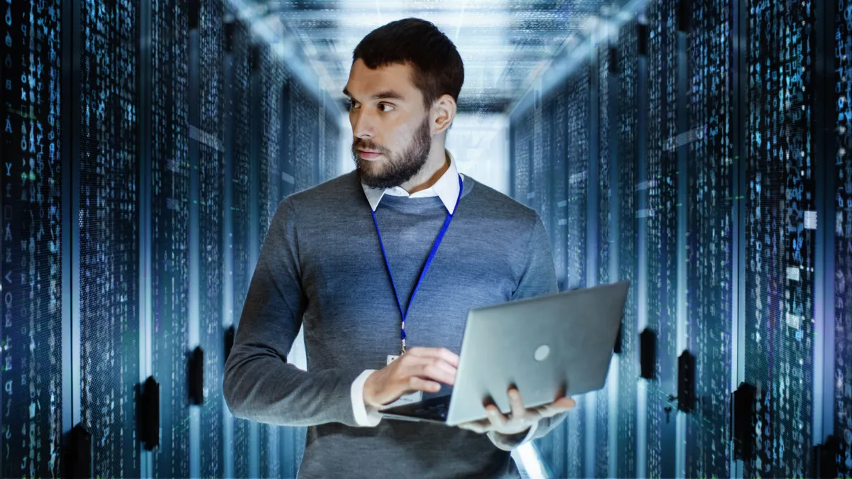 Male IT Server Technician Specialist Holds Laptop and Looking on Raining Script Code in Rack Sever Cabinet.