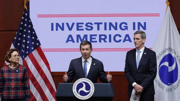 Man in suit at a podium in front of sign reading "Investing in America" with another man on the right and a woman on the left.