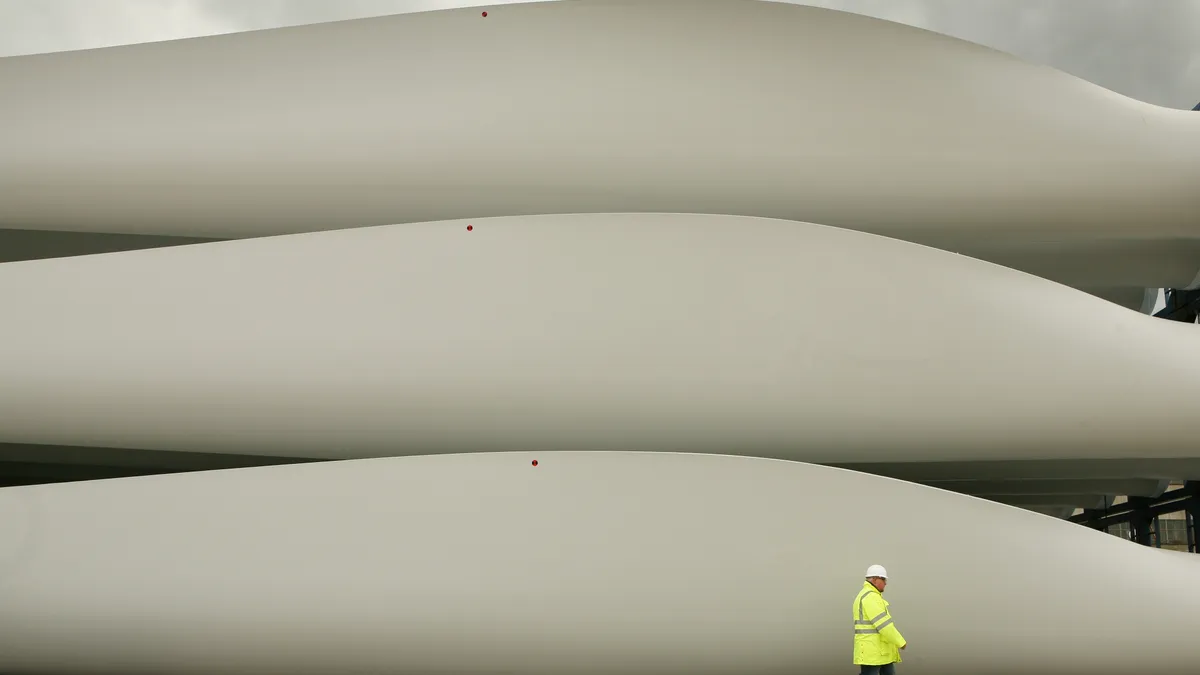 A worker wearing a high-visibility jacket walks past massive, off-white stored wind turbine blades at a shipyard.