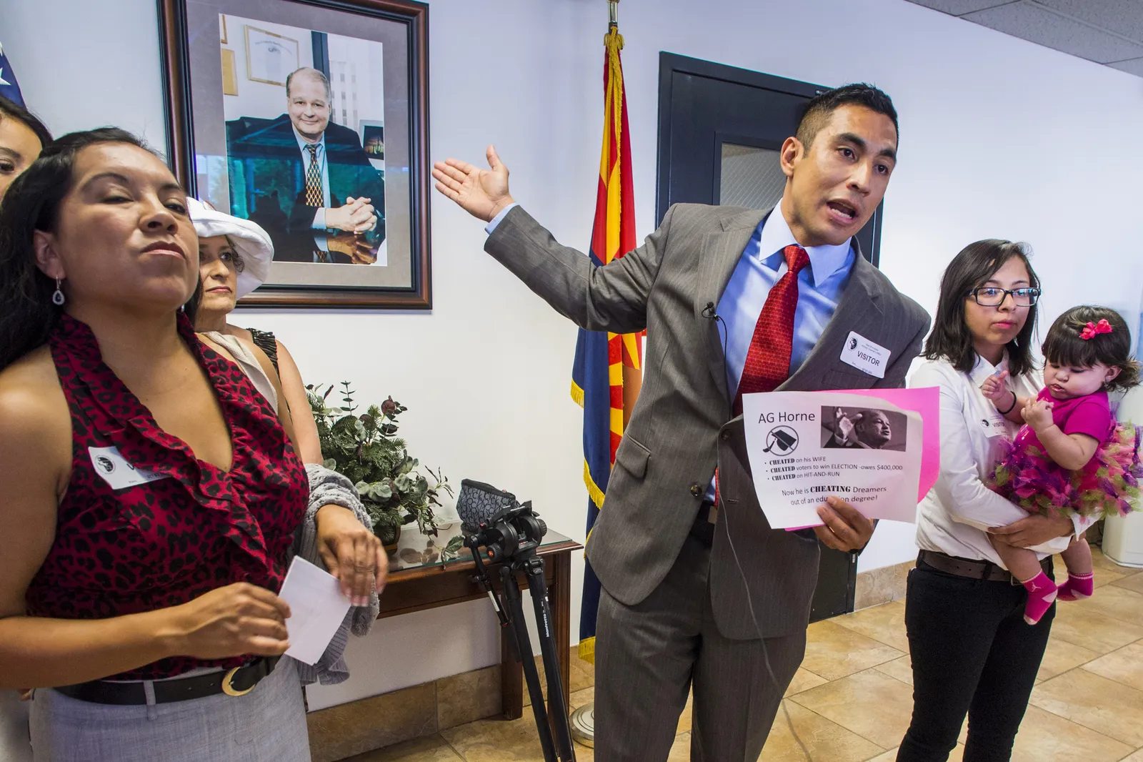 Several people gather in front o a portrait and hold a sign that reads, " Now he is cheating dreamers out of an education degree."