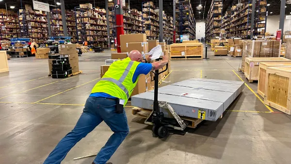 A person wearing a neon green worker vest pushes a large flat shipment on a pallet jack into a warehouse with rows of supply.