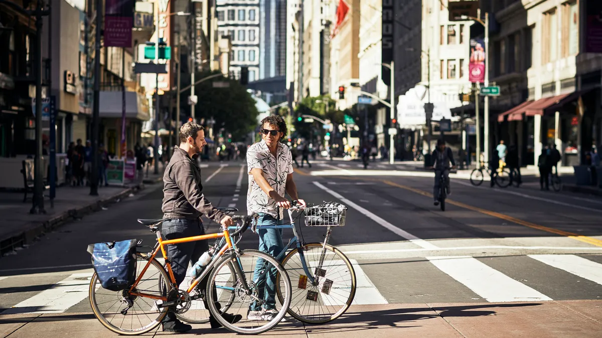 Two people rolling bikes next to them walk across a crosswalk on a city street