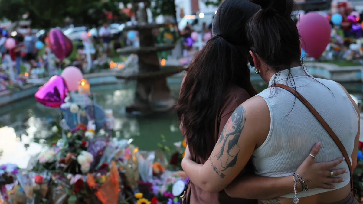 Two people standing and hugging outside with backs toward camera look at flowers, balloons and candles.