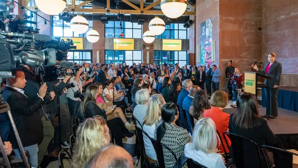 A woman stands at a podium in front of a large crows and TV cameras.