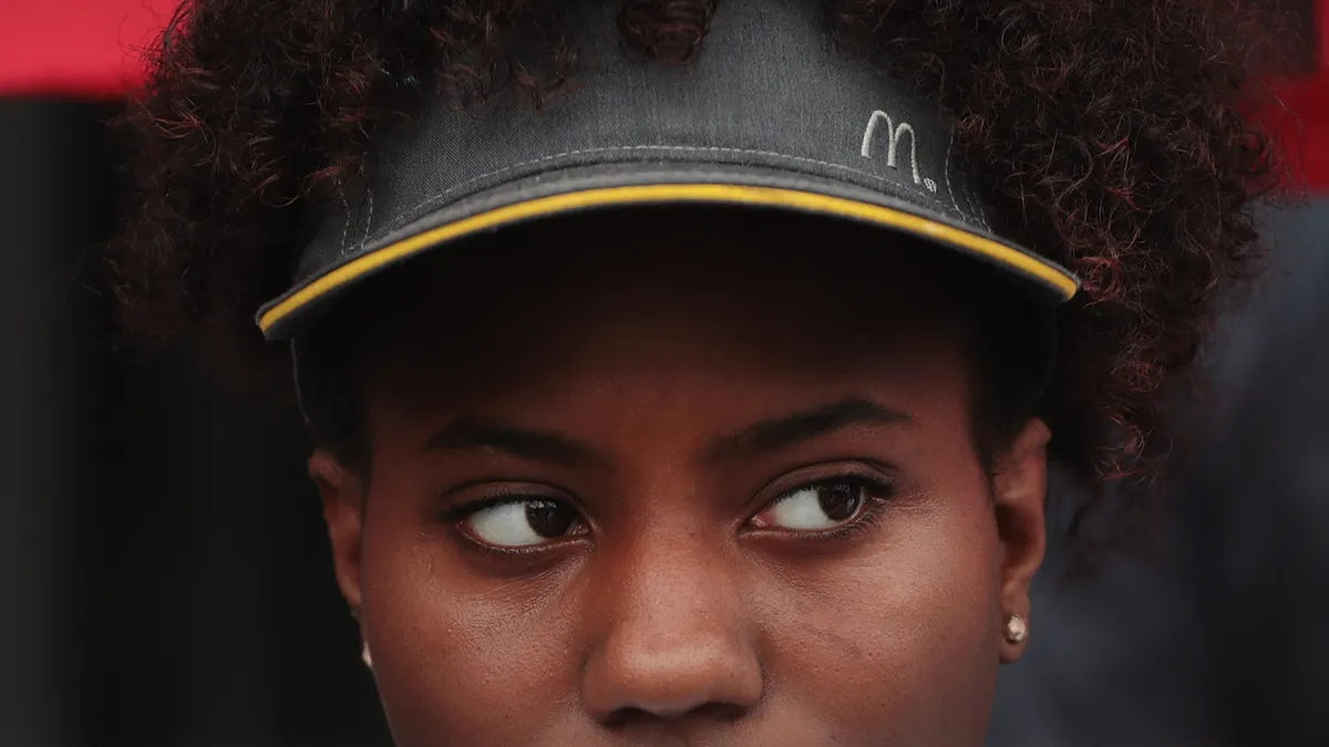 A Black worker wearing a McDonald's visor, with expresive eyes; they hold up an sign with the image of Ronald McDonald. It says, "Times Up, Clown."