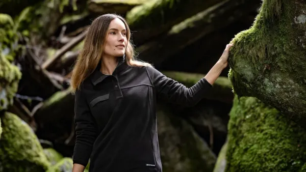 A woman is in a forest touching a mossy tree wearing a black fleece shirt