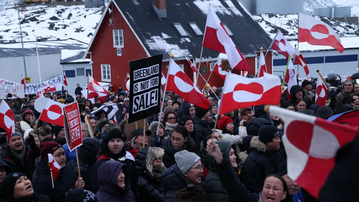 People wave the Greenland flag and hold signs during a protest. The signs read "Greenland is not for sale!"