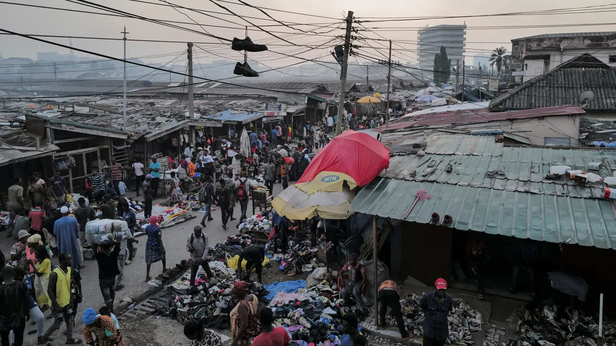 People walk in a crowded marketplace filled with bundles of clothes and covered with umbrellas and metal roofing.