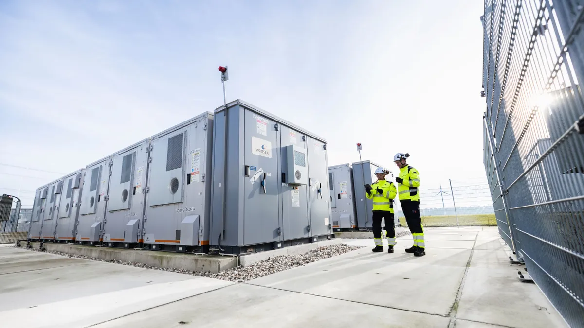 Two engineers in safety gear inspect large battery storage units at an outdoor facility with wind turbines in the background.