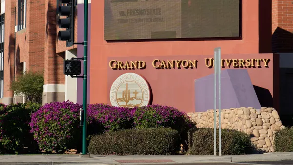 A large "Grand Canyon University" sign is adhered to a red stucco wall; above the sign is a large screen monitor.