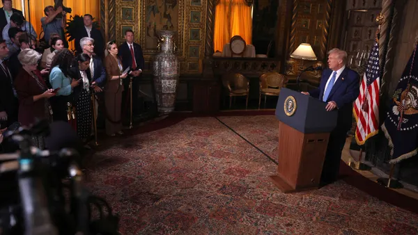 At right, President Trump stands at a podium in a room at Mar-a-Lago Resort, facing reporters, who stand behind ropes to the left.
