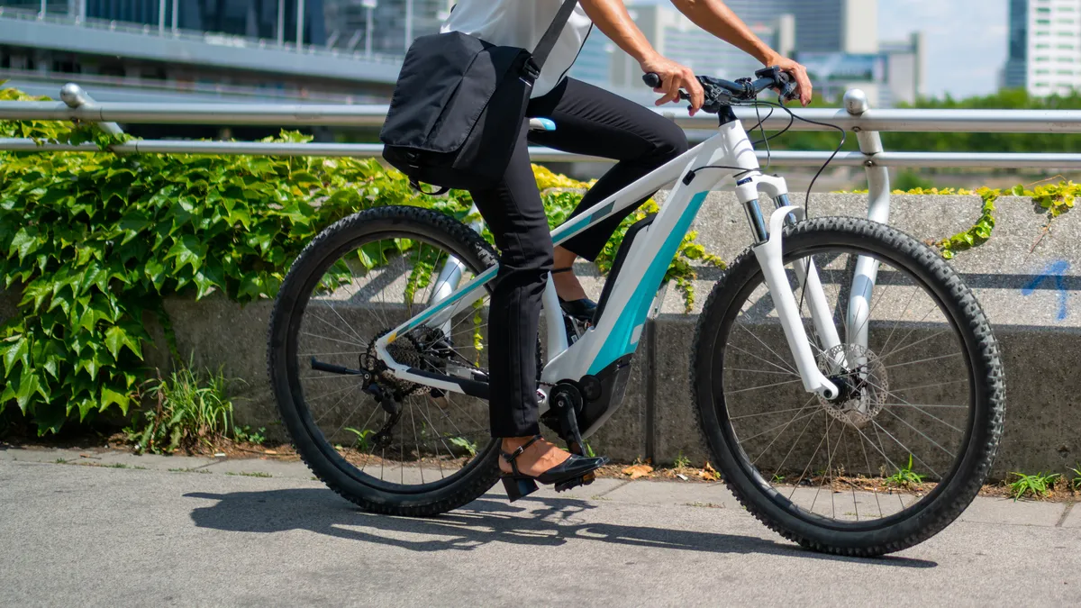 Business woman cycling on electric bicycle outdoors on sunny summer day in european city, office buildings blurred in background.