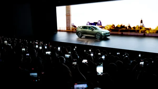 A Volvo EX60 is shown on stage during a reveal event, with the lettering EX 60 on a screen behind it.