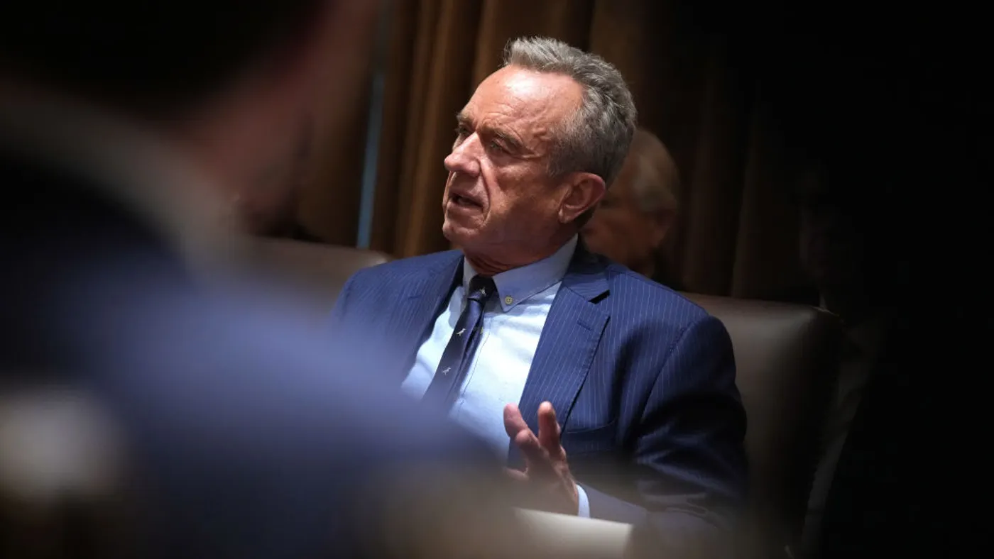 A person in a blue suit gestures while seated at a conference table.
