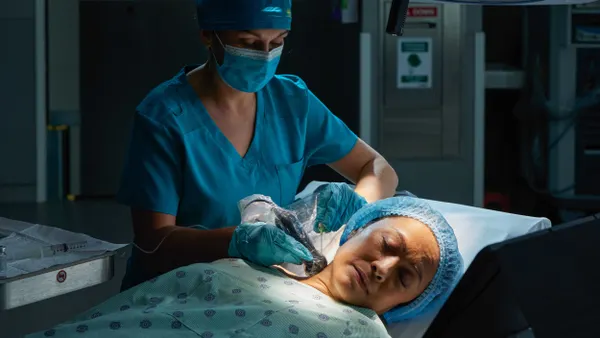 A pereson wearing a blue mask and scrubs holds an ultrasound probe next to a person's neck.