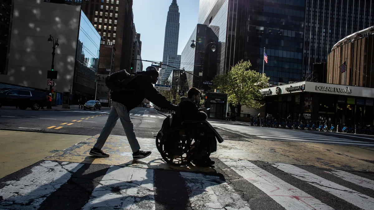 A man in a wheelchair crosses a street in Manhattan pushed by another man.