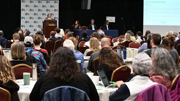 One person stands at a podium, with three seated, in front of a crowd of attendes at a MassRecycle event.