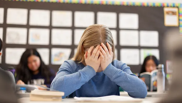 A student sitting at a desk in a classroom as their hands over their face.