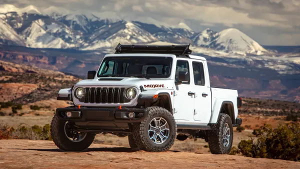 A white 2024 Jeep Wrangler parked on dirt terrain with snow-capped mountains in the background.