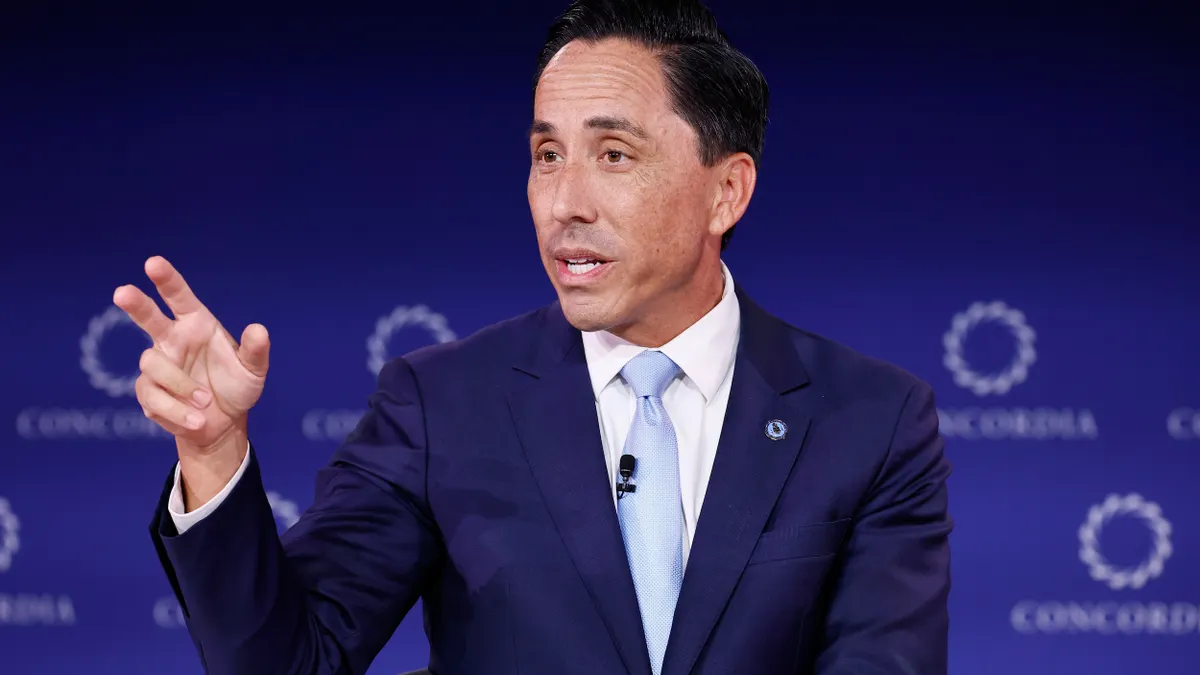 A person wearing a suit gestures with their hand while sitting in a chair in front of a blue background with Concordia labels.