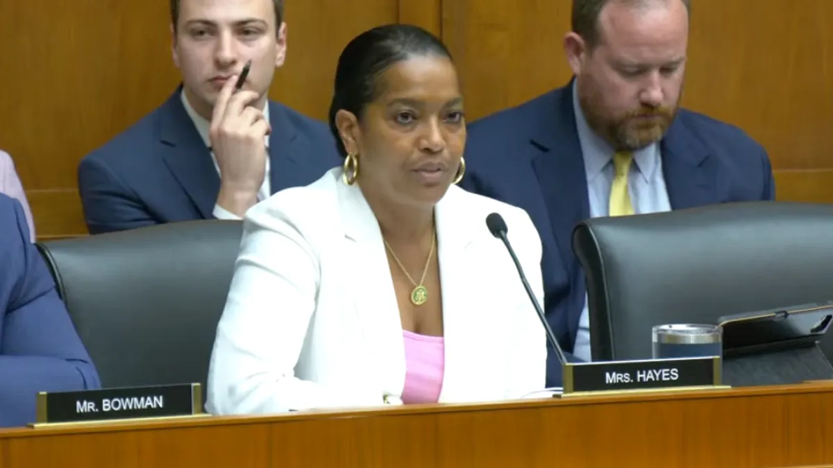 An adult sits at a desk in a hearing room on Capitol Hill. The nameplate in front of the person reads Mrs. Hayes.