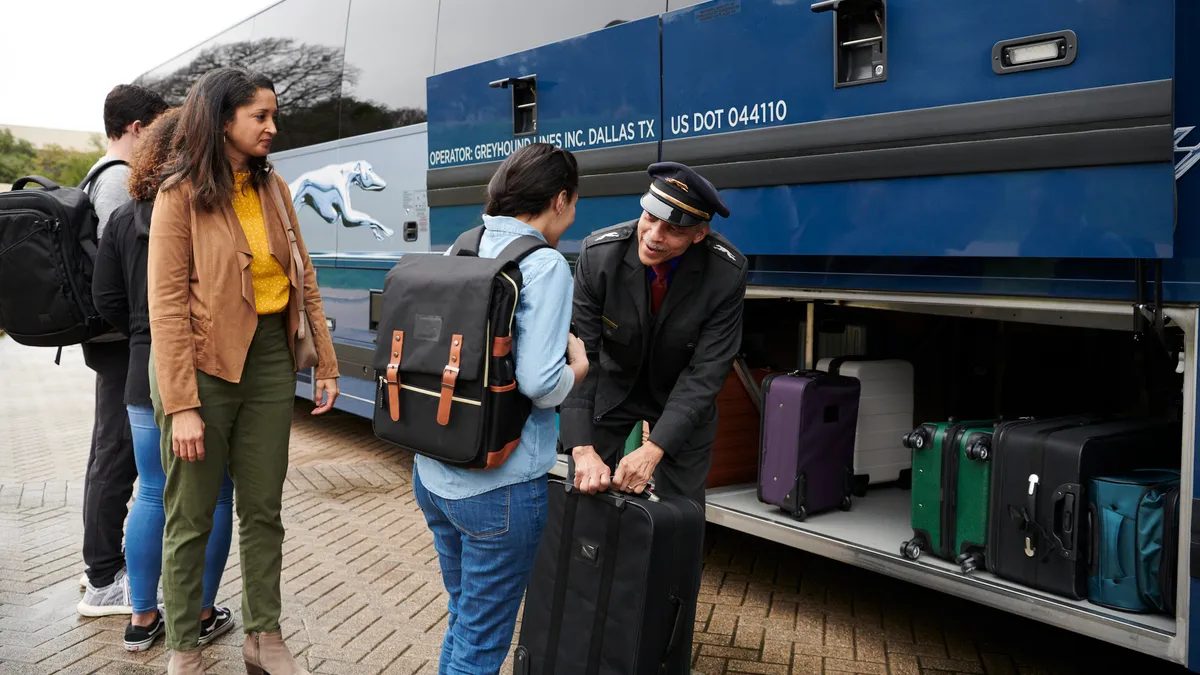 Four people are in line to board a blue and gray Greyhound bus, with one person handing her luggage to the driver standing in front of the open luggage door of the bus.