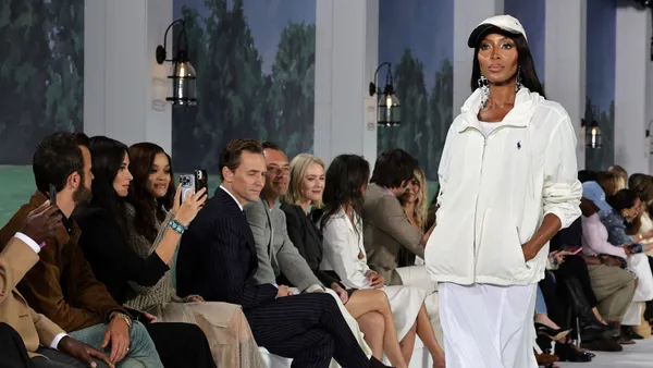 A model, Naomi Campbell, walks down a runway wearing a white jacket as viewers watch from the side.