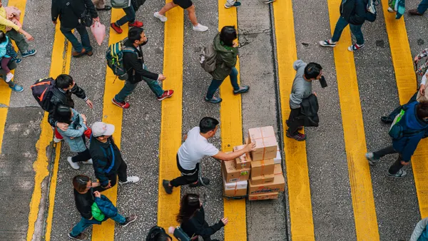 Overhead view of pedestrians and a delivery worker pushing stacked boxes across bright yellow crosswalk lines in busy Hong Kong, capturing urban movement, daily commute and street life energy.