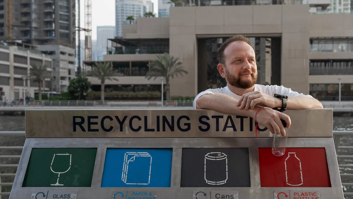 Sergey Shayakhmetov stands behind a row of recycling bins with a recyclable bottle in his hand.