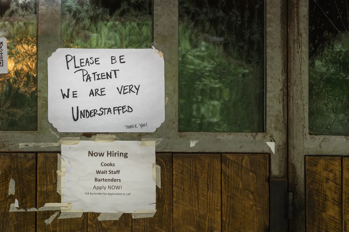 An image of two signs, one that says "Please be patient we are understaffed" and "Now hiring cooks, wait staff, bartenders. Apply now."