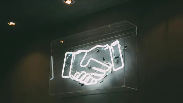 Illuminated neon sign of a handshake inside a transparent case against a dark wall. The soft glow suggests themes of partnership and agreement.
