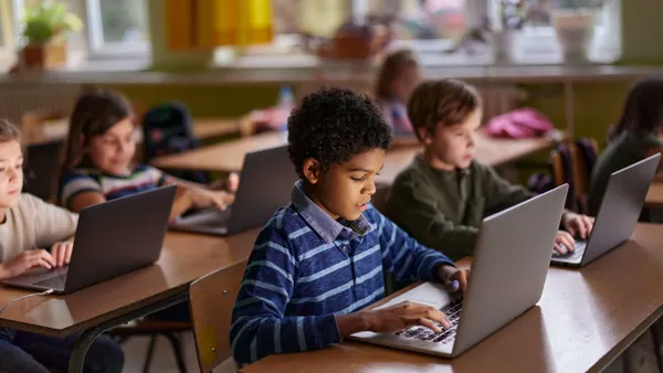 Elementary students sit next to each other in rows of desks while working on laptop computers.