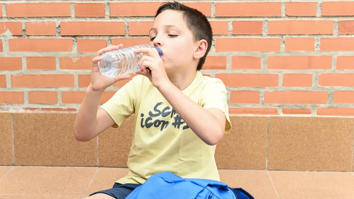 A child sitting next to a backpack in front of a brick wall drinks from a plastic water bottle.