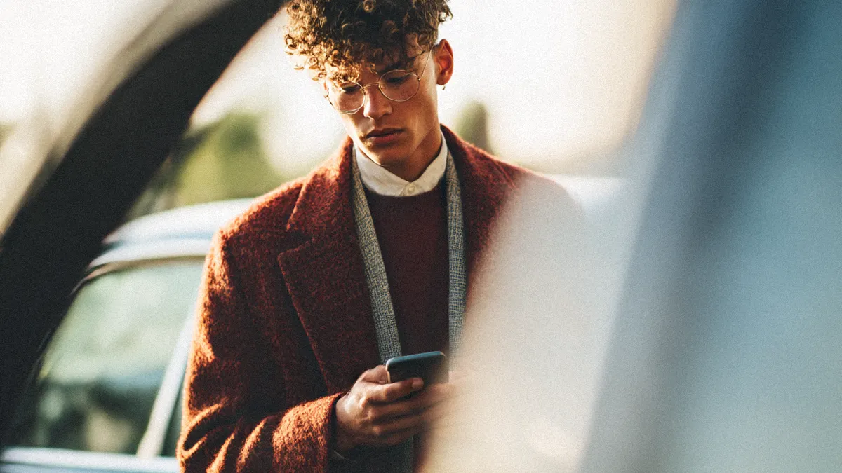 A young man with curly hair and glasses wears a textured rust-colored coat and sweater while looking down at a smartphone outdoors near a vehicle.