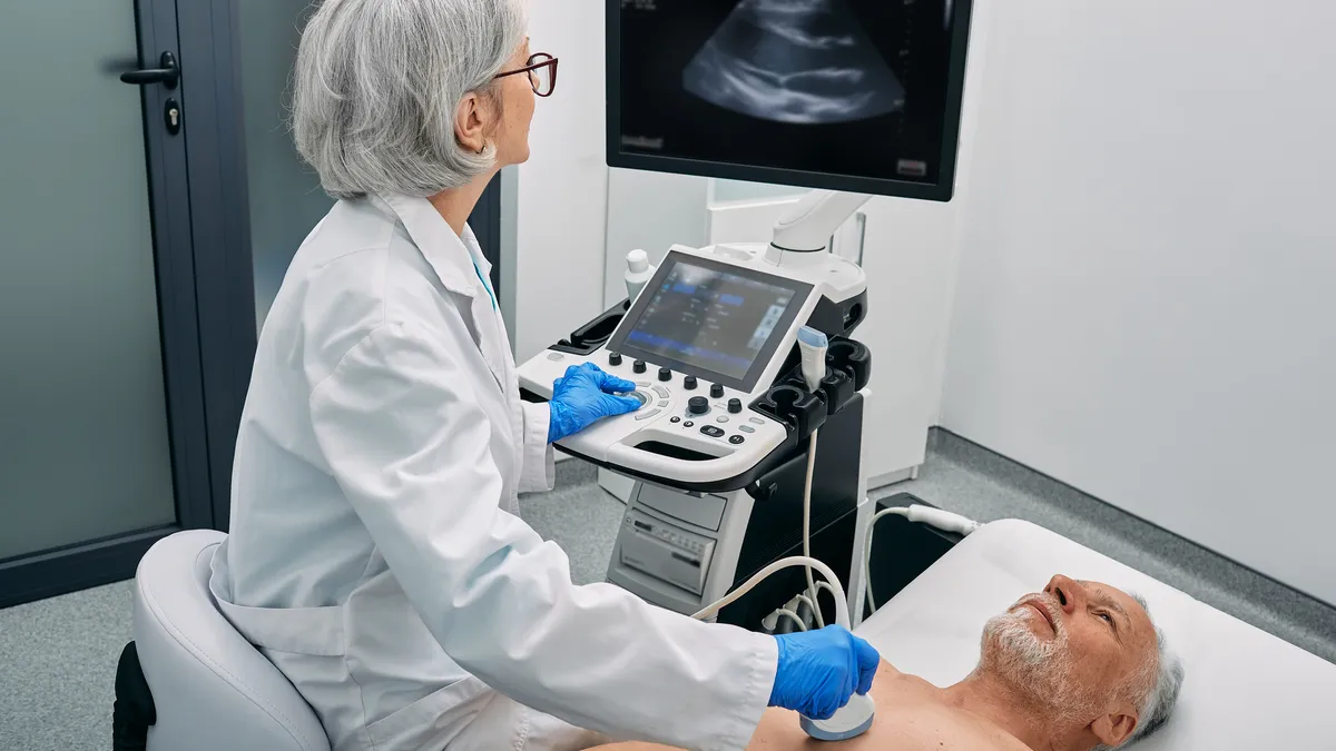 A person looks at a black and white image on a screen as they hold an ultrasound device to another person's chest.