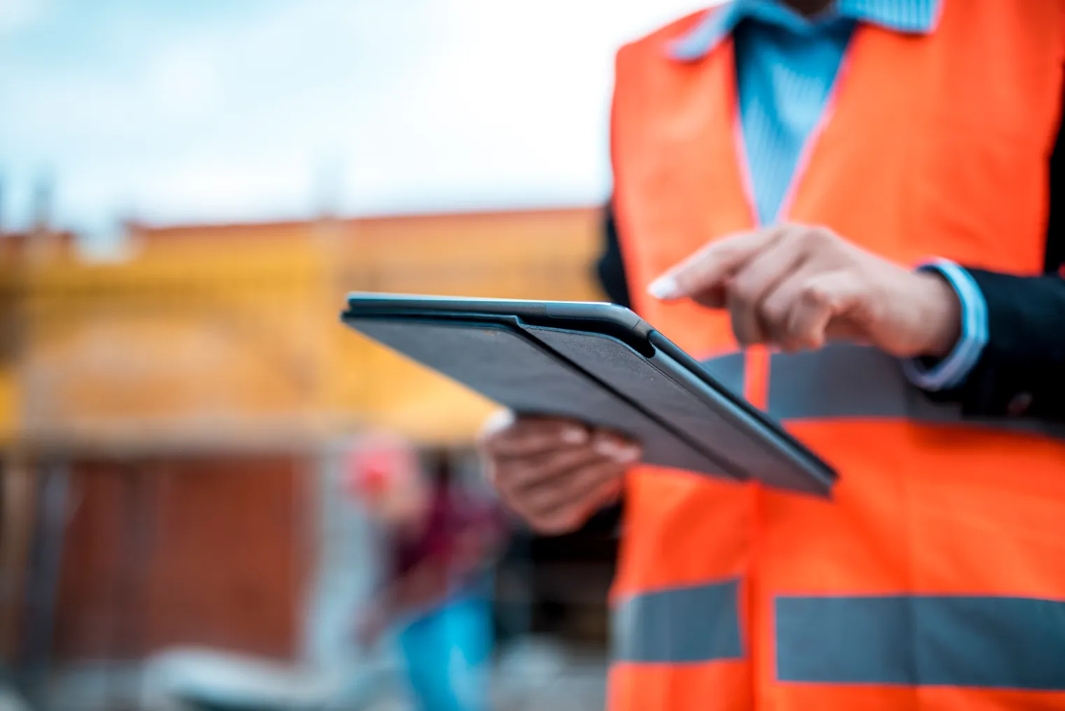 A man in a suit and an orange safety vest uses a tablet on a construction site. The user is in the foreground and clear, the background is blurred.