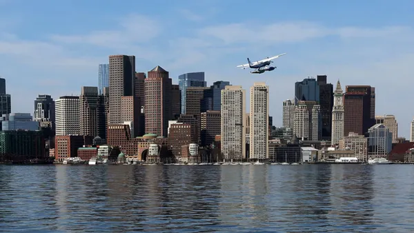 Boston Harbor is shown, depicting water in the foreground, tall buildings behind and blue sky in the background. A small plane flies above the water.