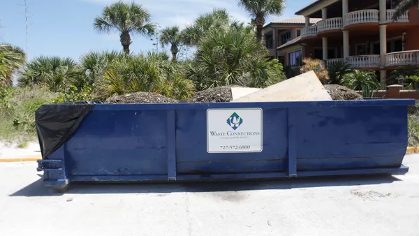 A large garbage container with the Waste Connections Logo sits on a concrete lot with palm trees and other vegetation behind it.